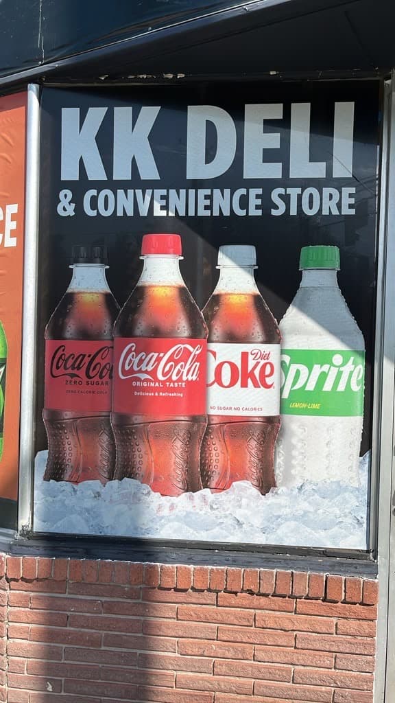 Storefront window graphic reading KK Deli & Convenience Store above bottles of soda on ice.